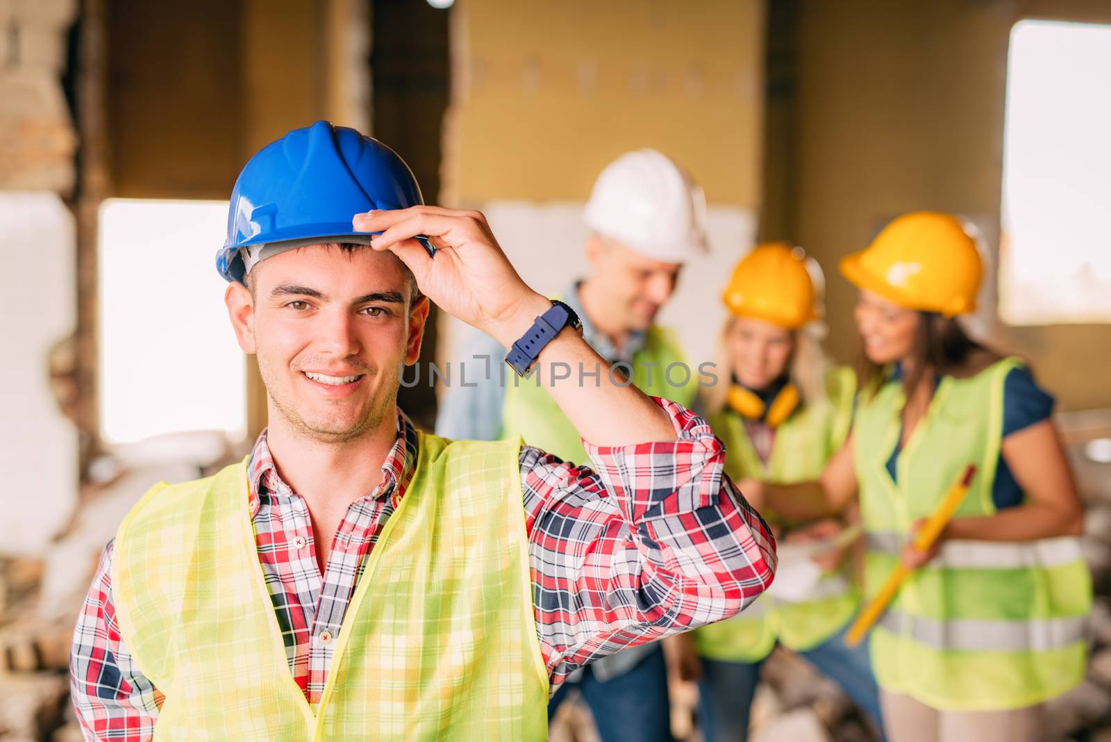 Smiling young electrician holding helmet and looking at camera. His architect colleagues review plan in background at a construction site.