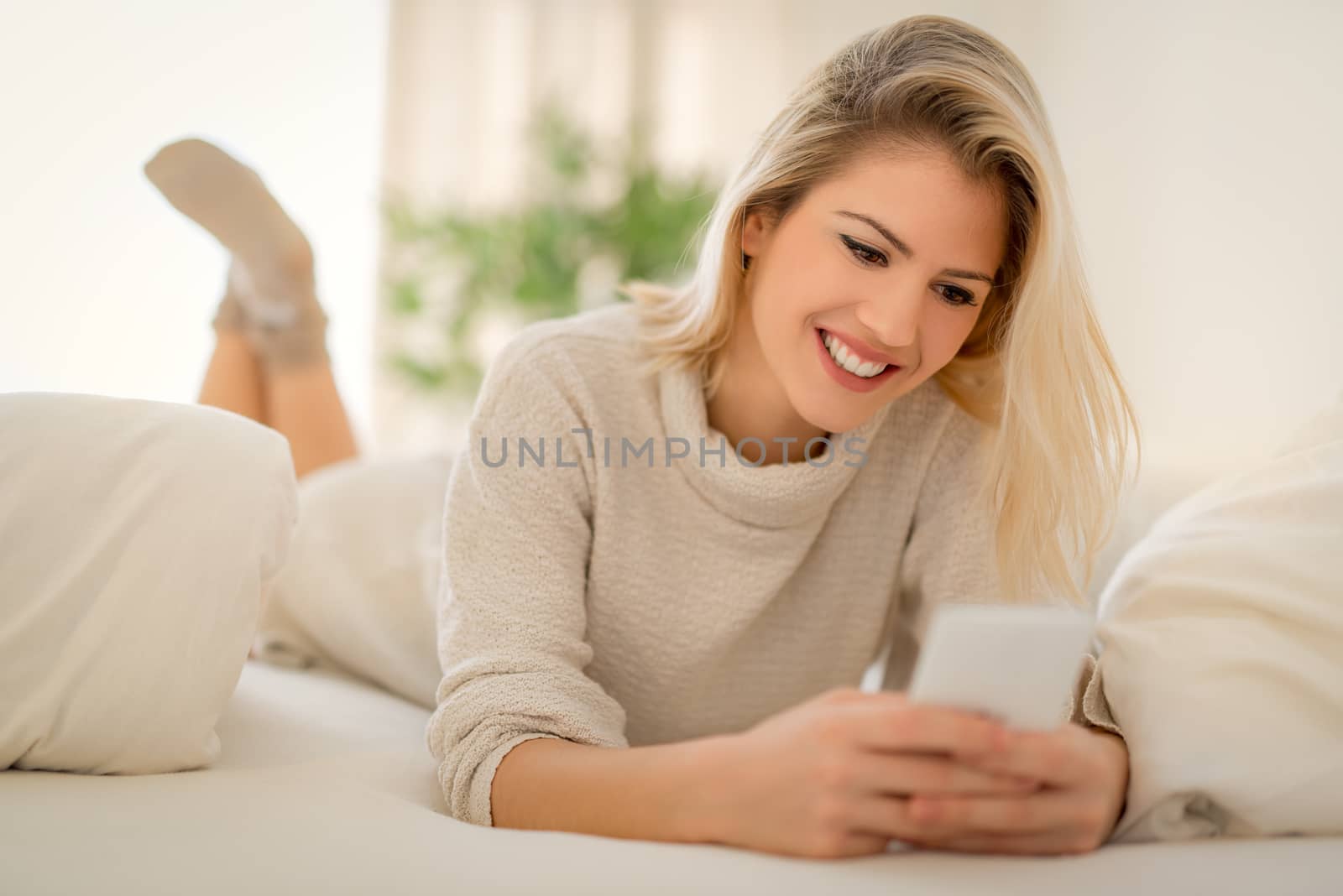 Beautiful young smiling woman relaxing in bed and typing message on smart phone.