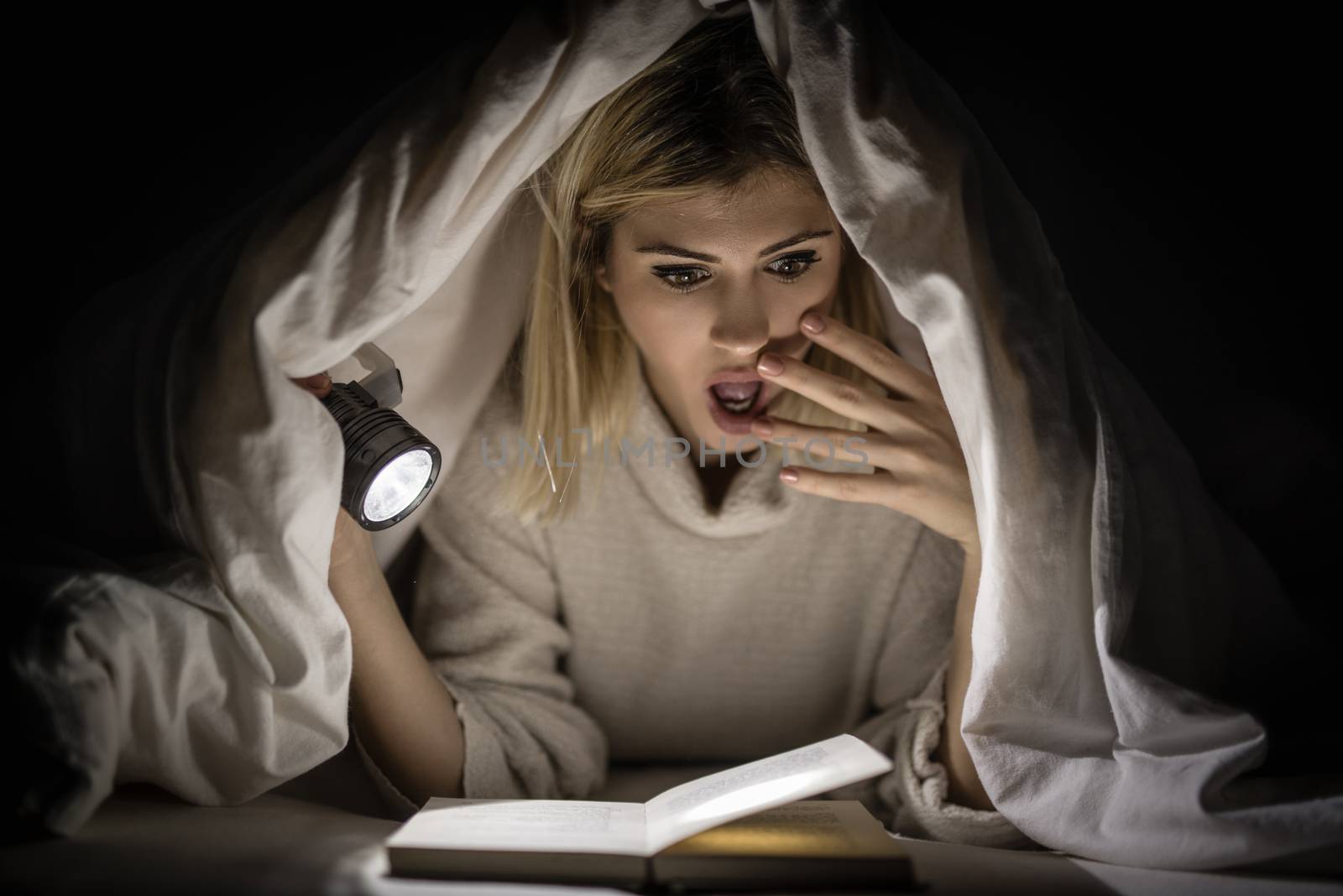 Beautiful young woman reading book under bed cover and holding a flashlight. 