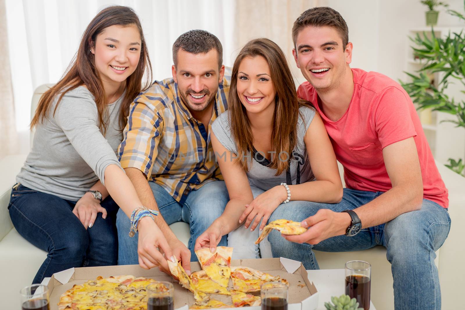 Four cheerful friends hanging out in an apartment. They eating pizza and looking at camera.