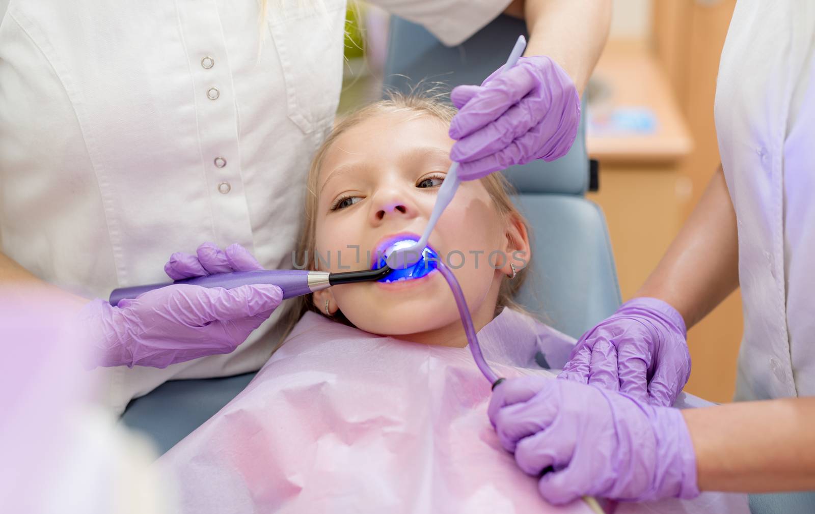 Beautiful little girl at visit in the dentist office. She is sitting on a chair and dentist finishing repair tooth with dental curing UV lamp. Selective focus.
