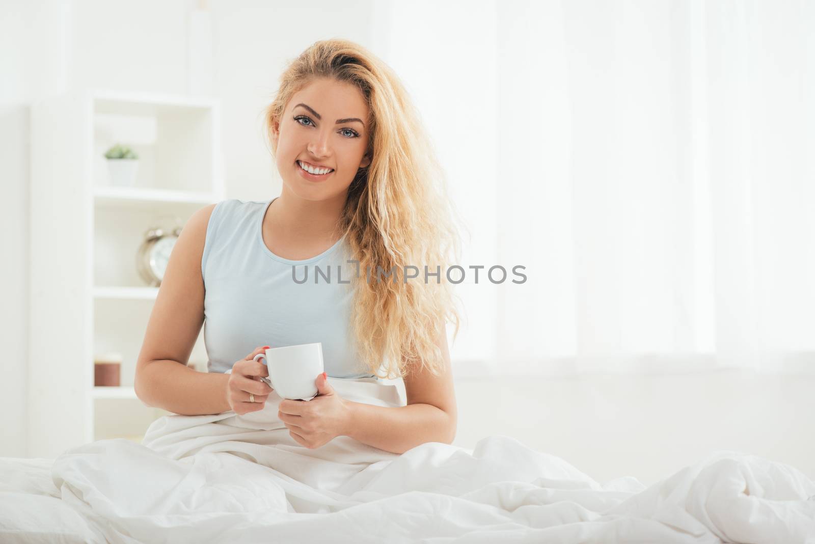 Young pretty girl relaxing in bed at morning. She is enjoying and drinking coffee. Looking at camera. 