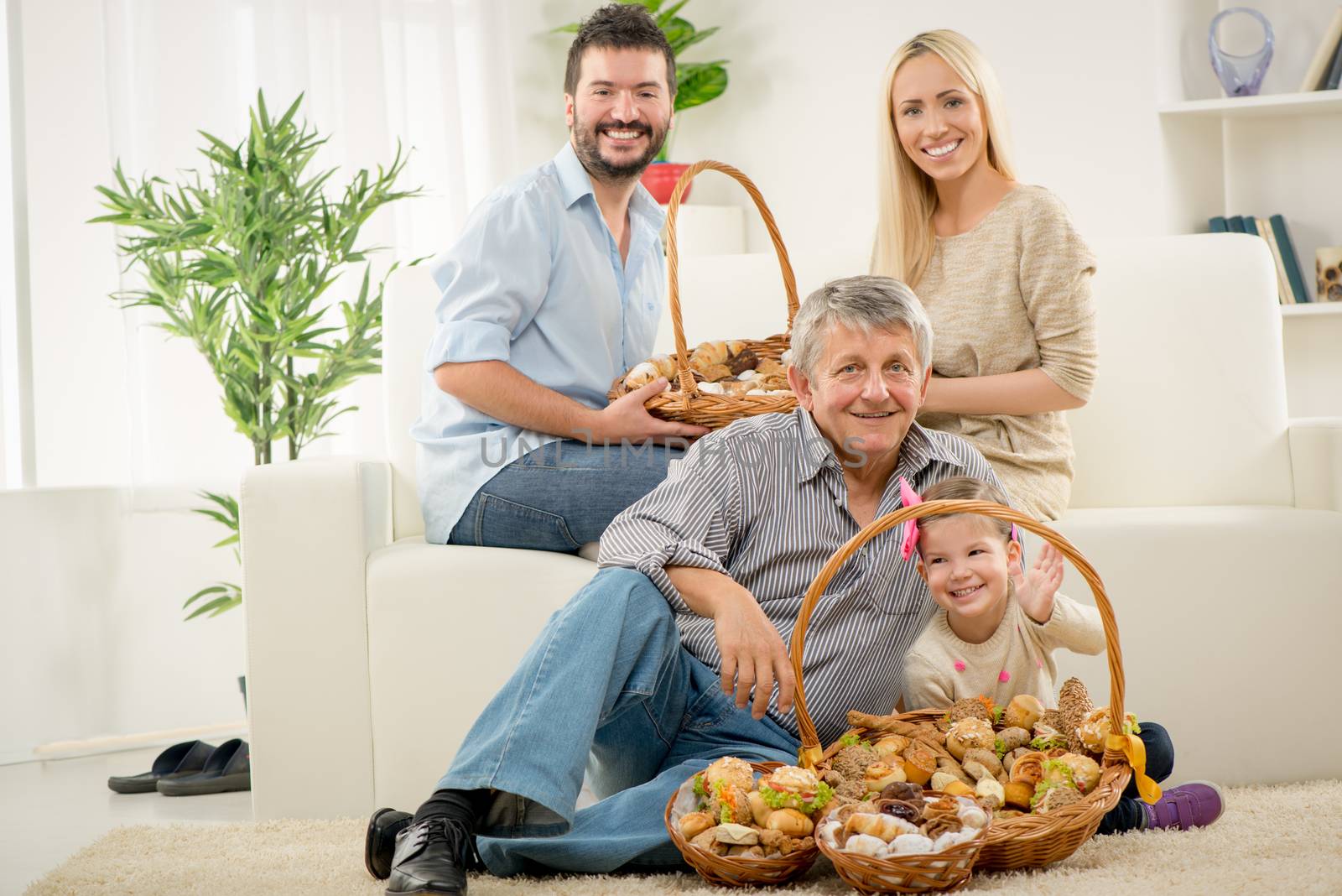 Senior man and a little girl are sitting on the living room floor. In front of them is basket with pastries, and behind them young parents are sitting on the couch. Looking at camera.