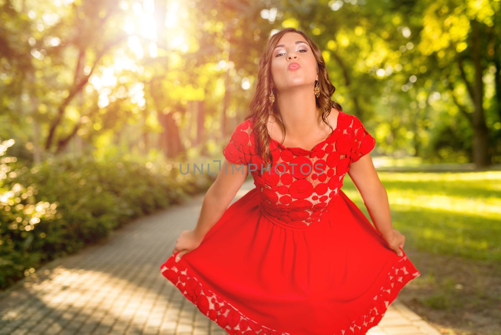 Young beautiful woman in red dress enjoy the sun in the park. She is send a kiss and looking at camera.