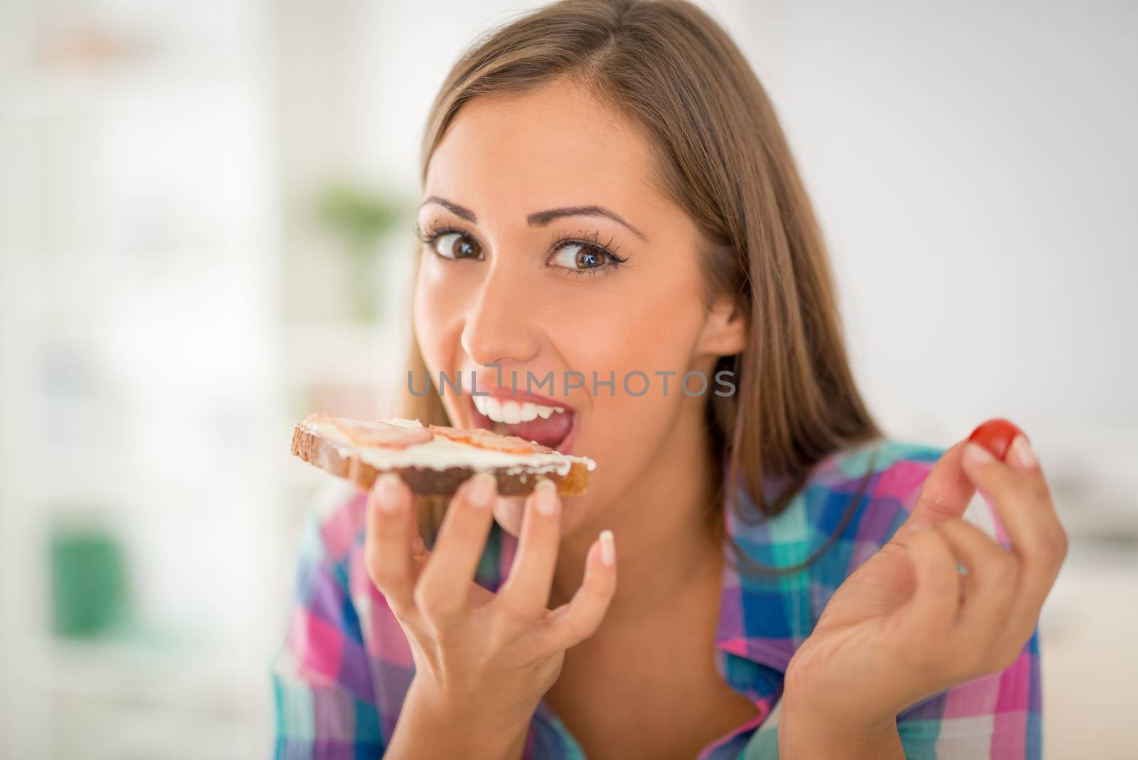 Beautiful young woman having breakfast in the domestic kitchen. She is eating fresh sandwich and drinking milk. Looking at camera.