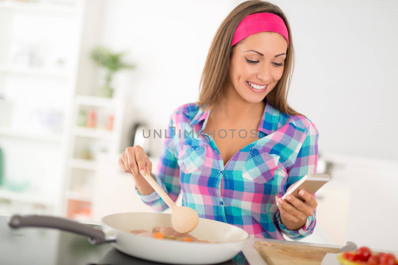 Beautiful young woman making healthy meal and using phone in the domestic kitchen.