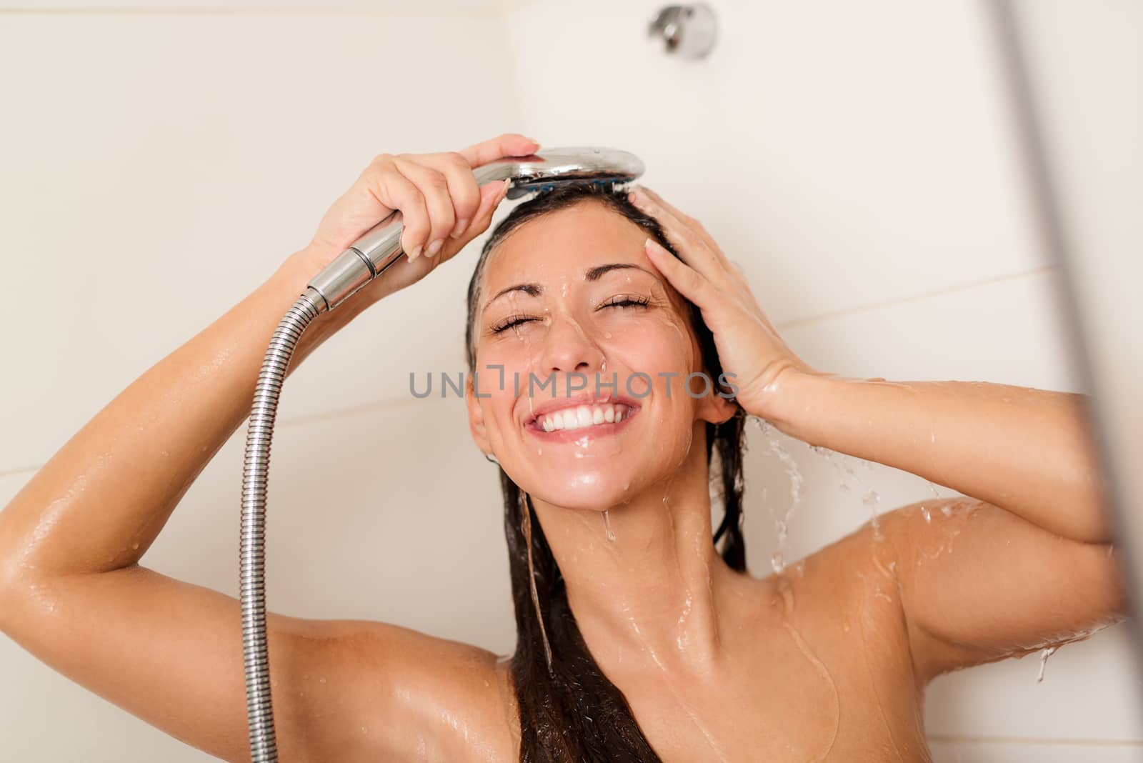 Happy young woman washing face and hair while showering under shower head.