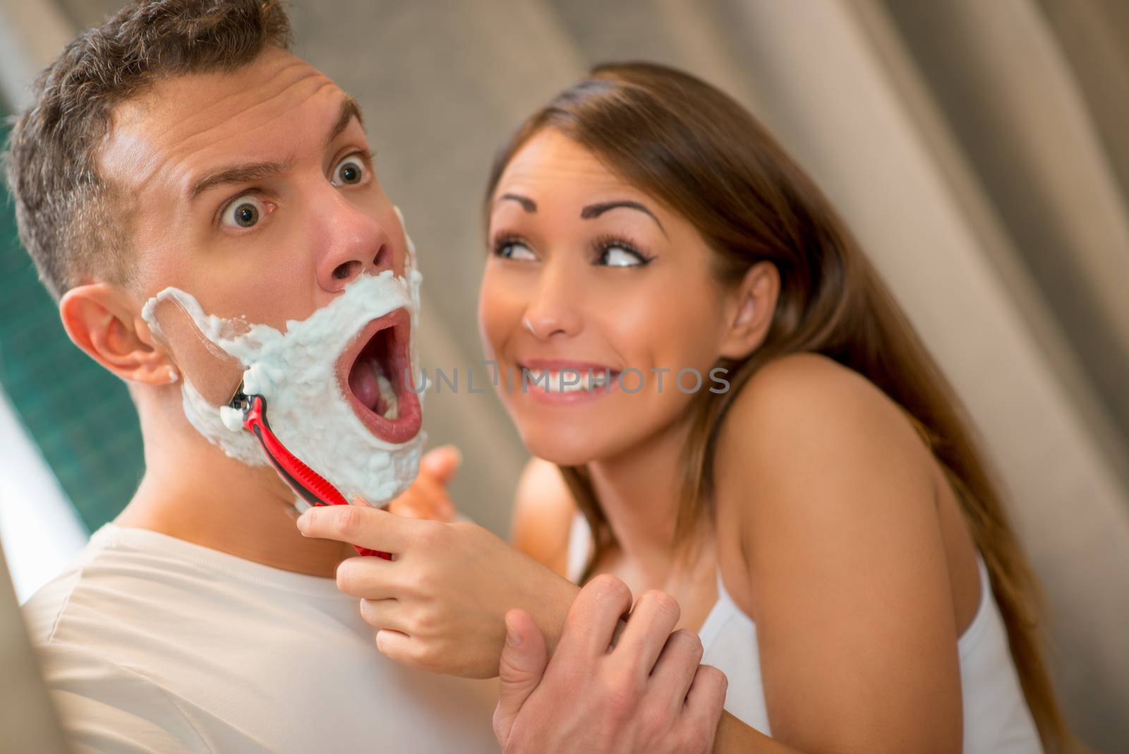 Beautiful young couple have fun in front of her bathroom mirror. Cheerful girl shaving her boyfriend. He is looking with scary face.