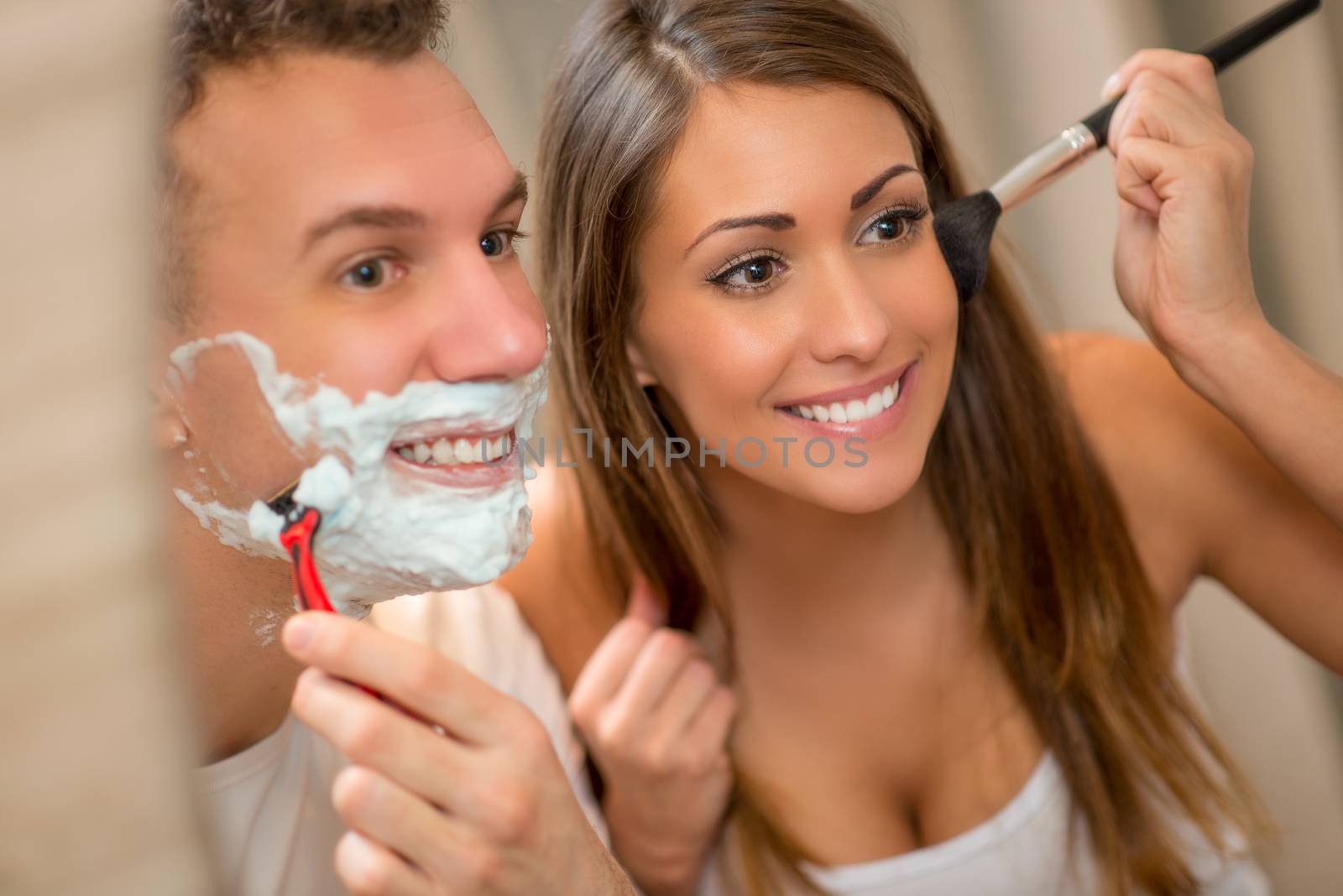 Portrait of a beautiful young happy couple having morning habits in front of their bathroom mirror. Man shaving and his girlfriend makeup. Selective focus.