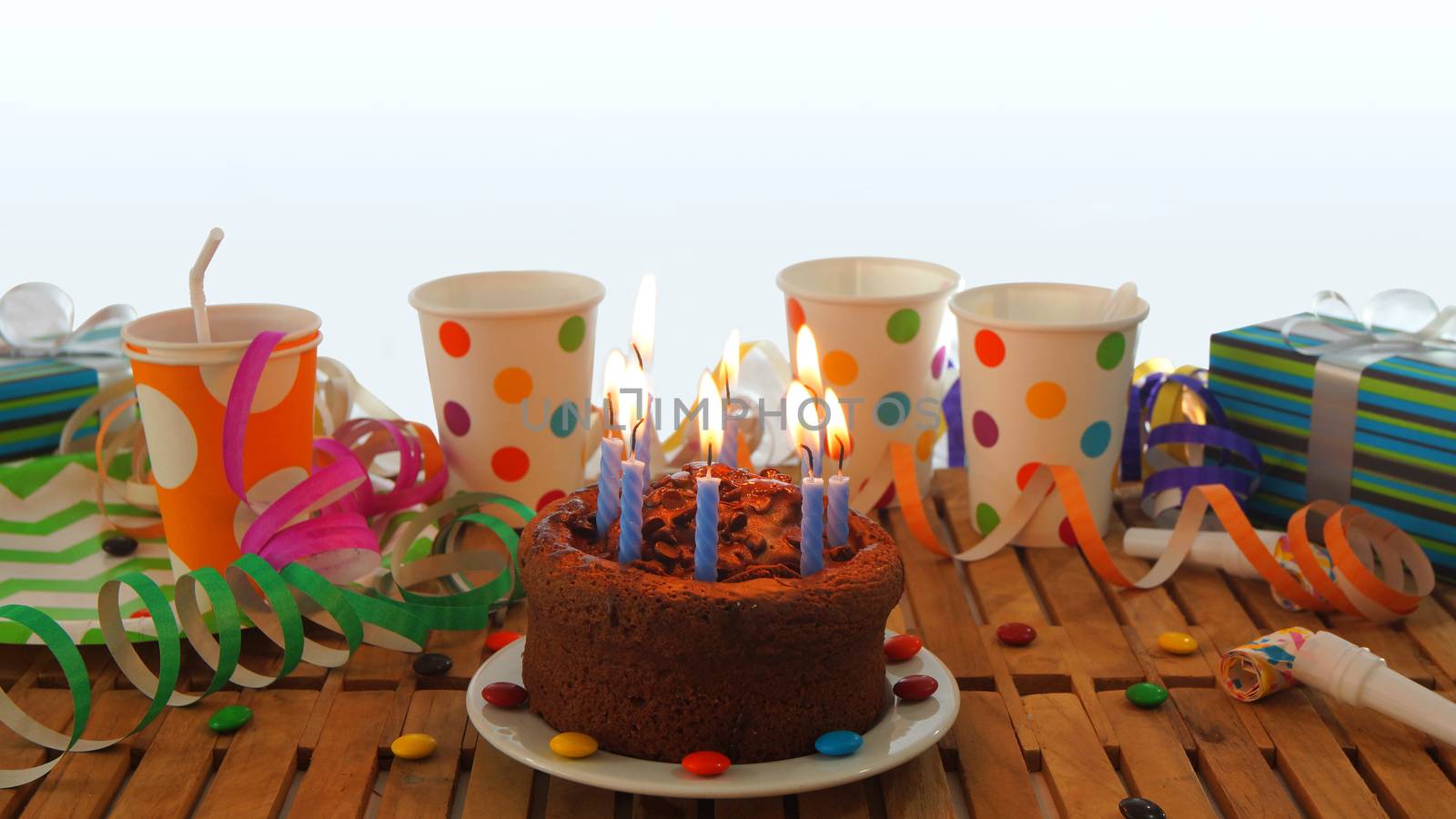 Chocolate birthday cake with a blue candles burning on rustic wooden table with background of colorful streamers, gifts, plastic cups with candies and white wall in the background