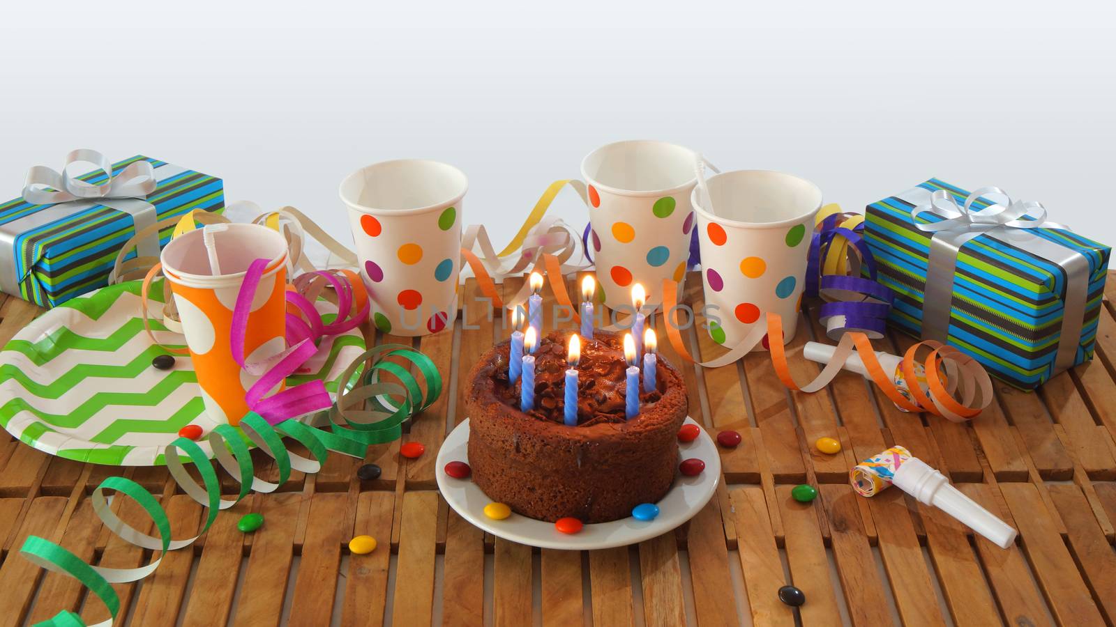 Chocolate birthday cake with a blue candles burning on rustic wooden table with background of colorful streamers, gifts, plastic cups with candies and white wall in the background