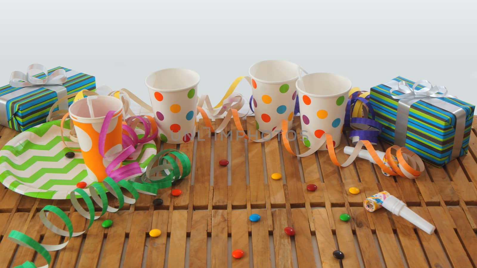 Rustic wooden table with colorful streamers, gifts, plastic cups, plastic plate, candies on white background. Party background