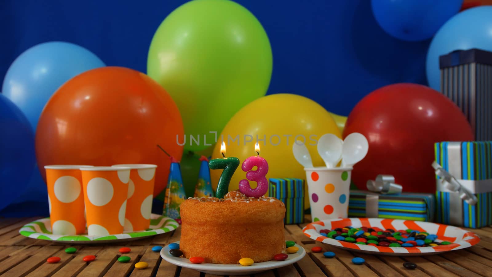 Birthday cake on rustic wooden table with background of colorful balloons, gifts, plastic cups and plastic plate with candies and blue wall in the background by alejomiranda