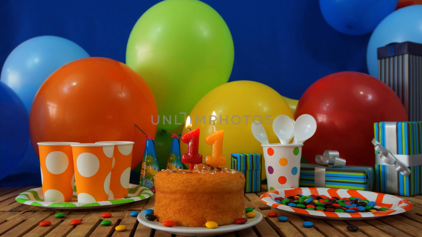 Birthday cake on rustic wooden table with background of colorful balloons, gifts, plastic cups and plastic plate with candies and blue wall in the background by alejomiranda