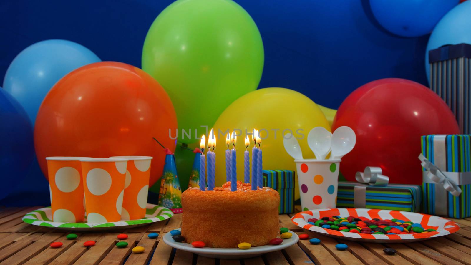 Birthday cake with a blue candles burning on rustic wooden table with background of colorful balloons, gifts, plastic cups and plastic plate with candies and blue wall in the background