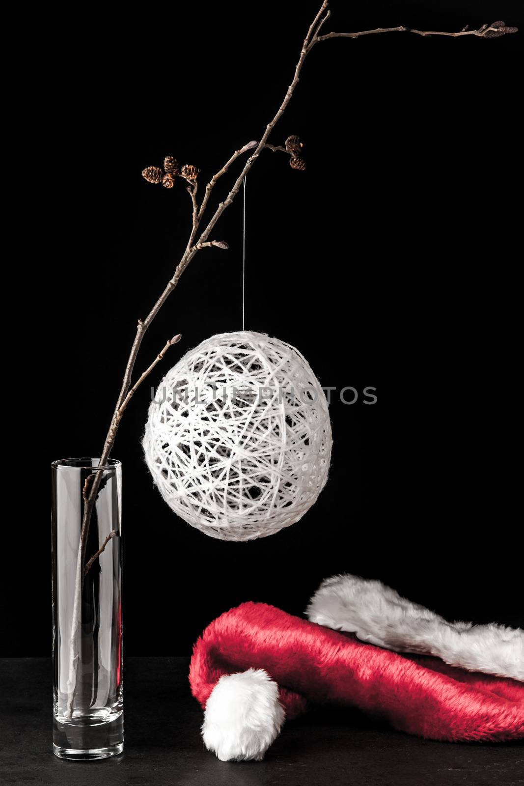 Alder branch in the glass with a white christmas ball and santa claus hat on the black stone table vertical