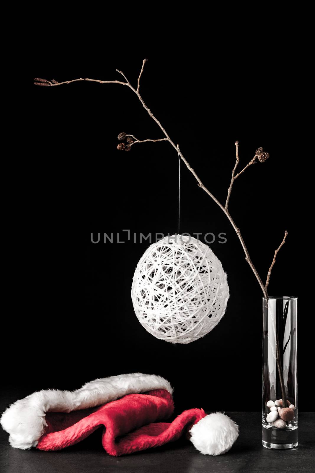 Alder branch in the glass with a white christmas ball and santa claus hat on the black stone table vertical