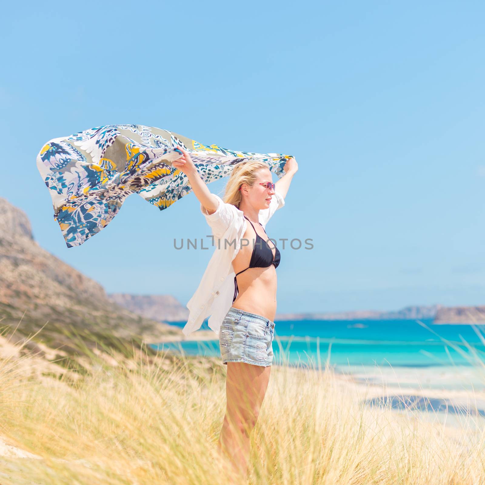 Relaxed woman, arms rised, holding colorful scarf, enjoying sun, freedom and life at beautiful beach. Young lady feeling free, relaxed and happy. Concept of vacations, freedom, joy and well being.