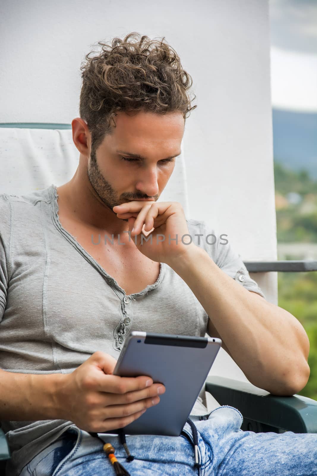 Handsome trendy man wearing t-shirt sitting and working, looking down at a tablet computer that he is holding, outdoor on house terrace