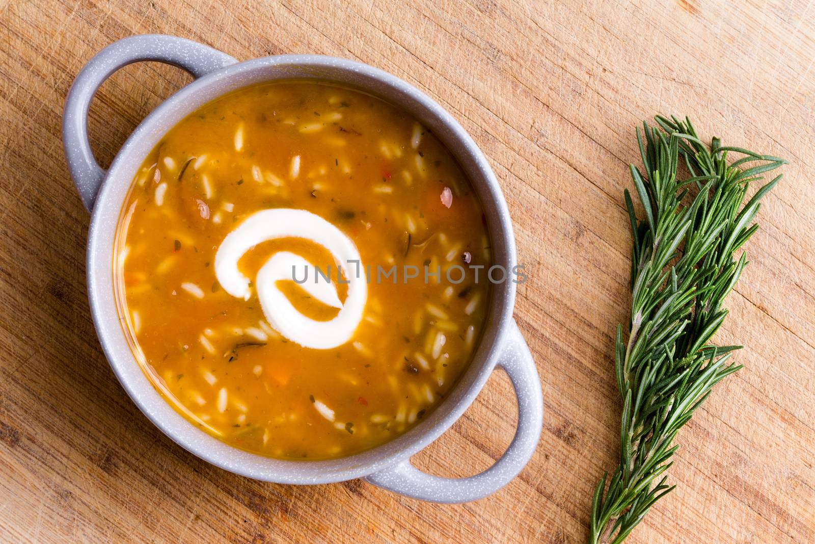 Cup of chicken broth with wild rice served with a twirl of sour cream ready to be garnished with fresh rosemary displayed alongside on a bamboo cutting board, overhead view