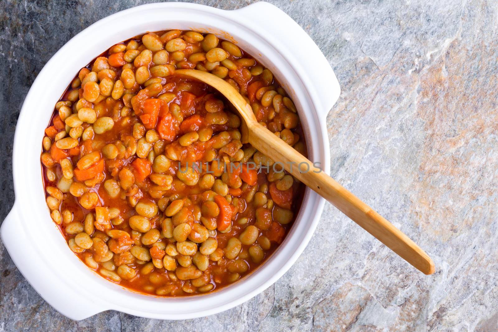 Baby lima bean casserole with carrots served in a white ceramic dish with a wooden serving spoon on a textured stone counter, overhead view with copy space