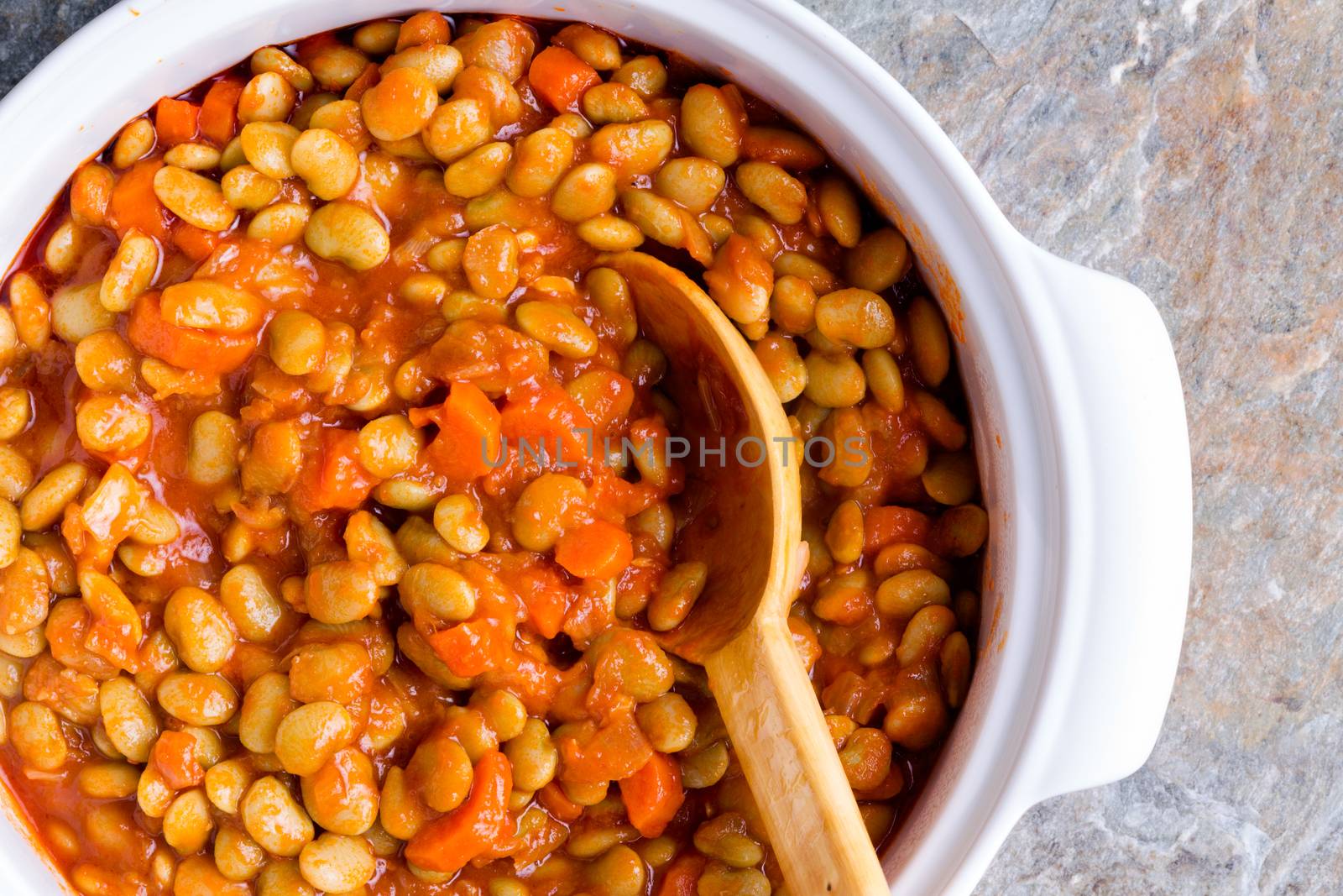 Delicious vegetarian lima bean and carrot vegetable casserole served in a white casserole dish with a wooden spoon viewed close up from above on a stone counter top