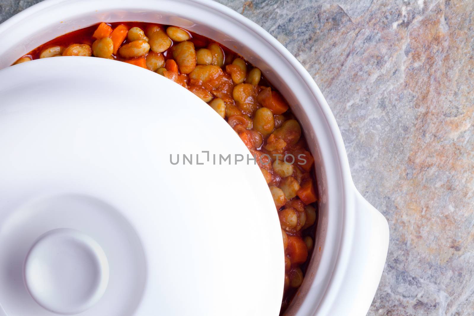 Delicious savory lima bean casserole with carrots cooling off in a white ceramic casserole dish with the lid moved aside to view the contents, closeup overhead view on a stone counter