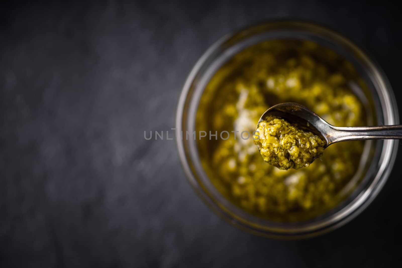 Pesto sauce in the metal spoon with blurred jar top view
