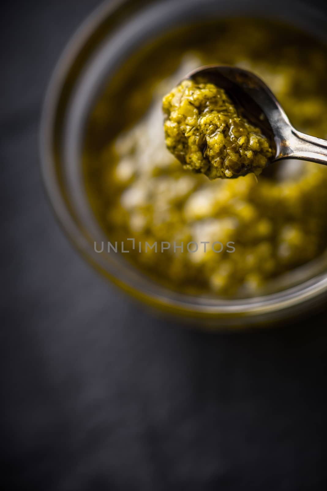 Pesto sauce in the metal spoon with blurred jar vertical by Deniskarpenkov
