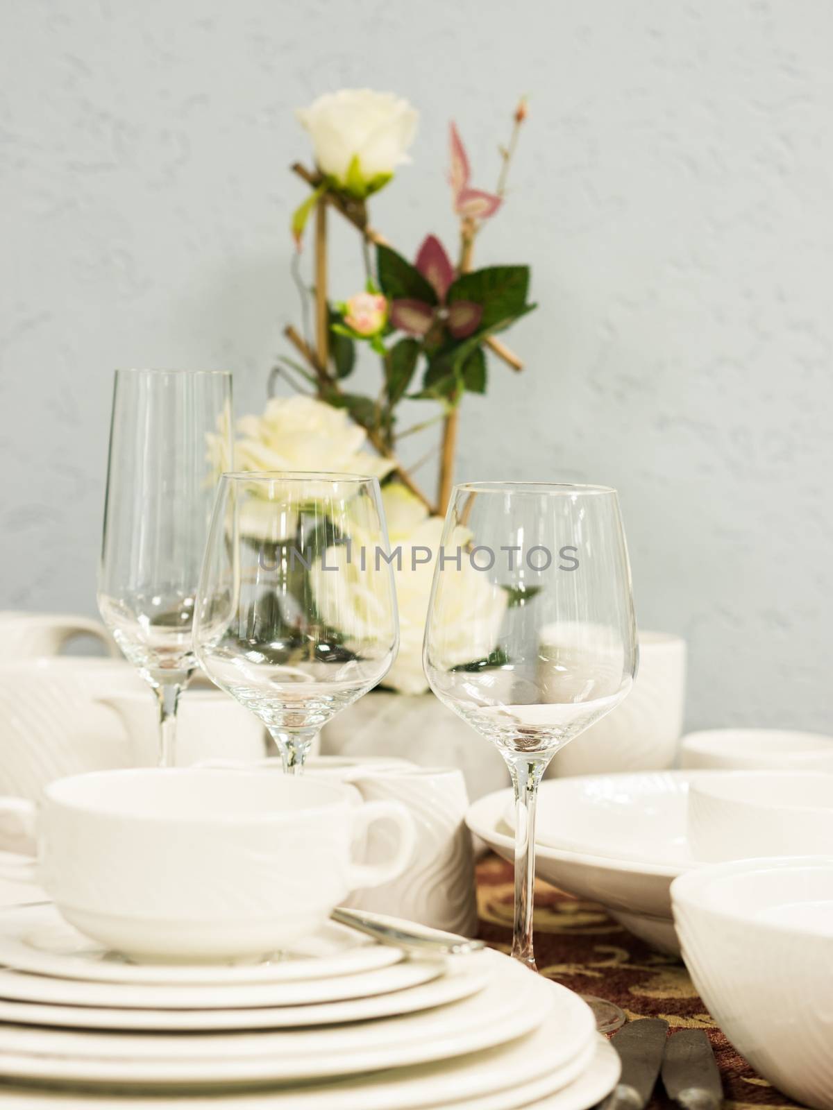 Set of new dishes on table with tablecloth. Stack of white plates and wine glasses with flowers on restaurant table. Vertical. Shallow DOF