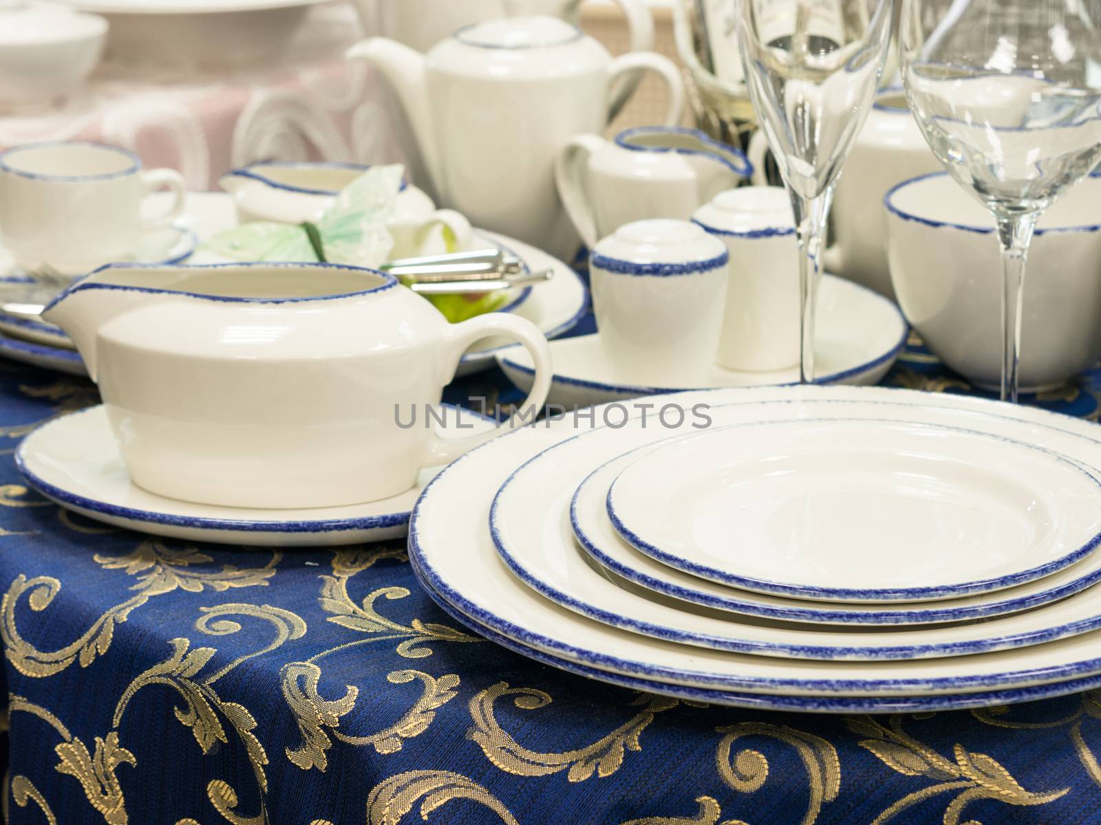 Set of new dishes on table with blue tablecloth. Stack of plates, saucer and wine glasses on restaurant table. Shallow DOF