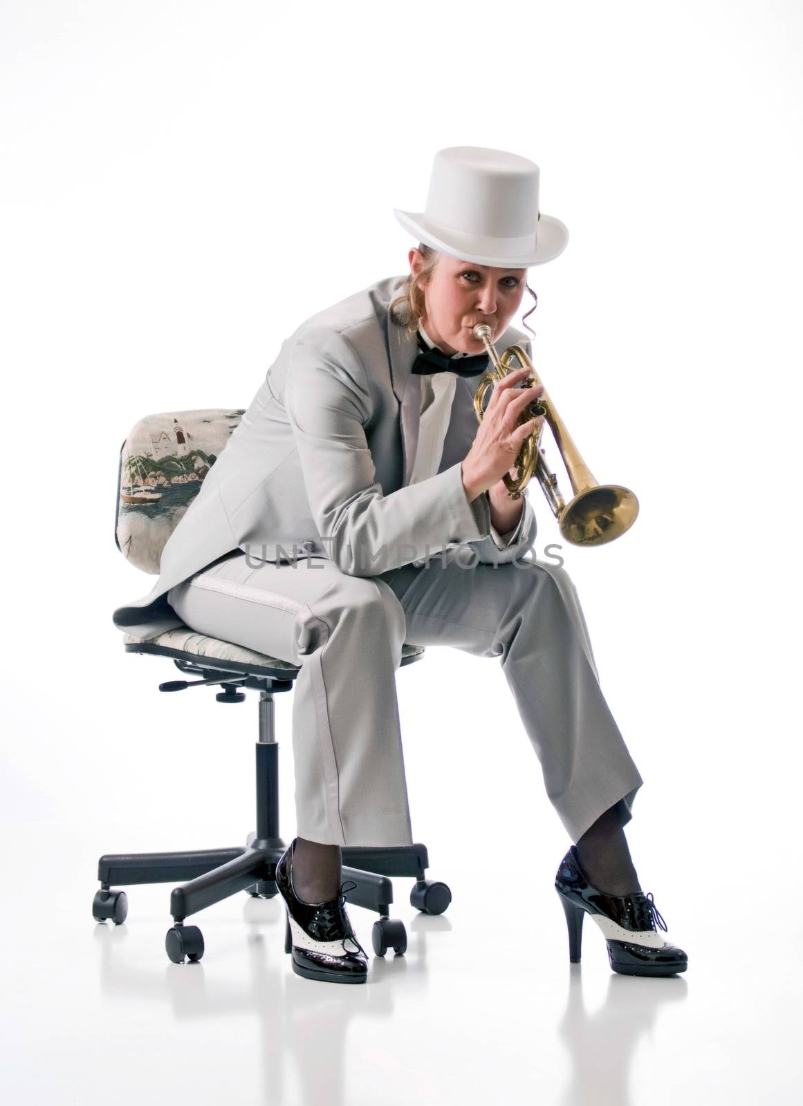 Pretty blonde woman in tuxedo and top-hat playing a trumpet while seated on a stool.