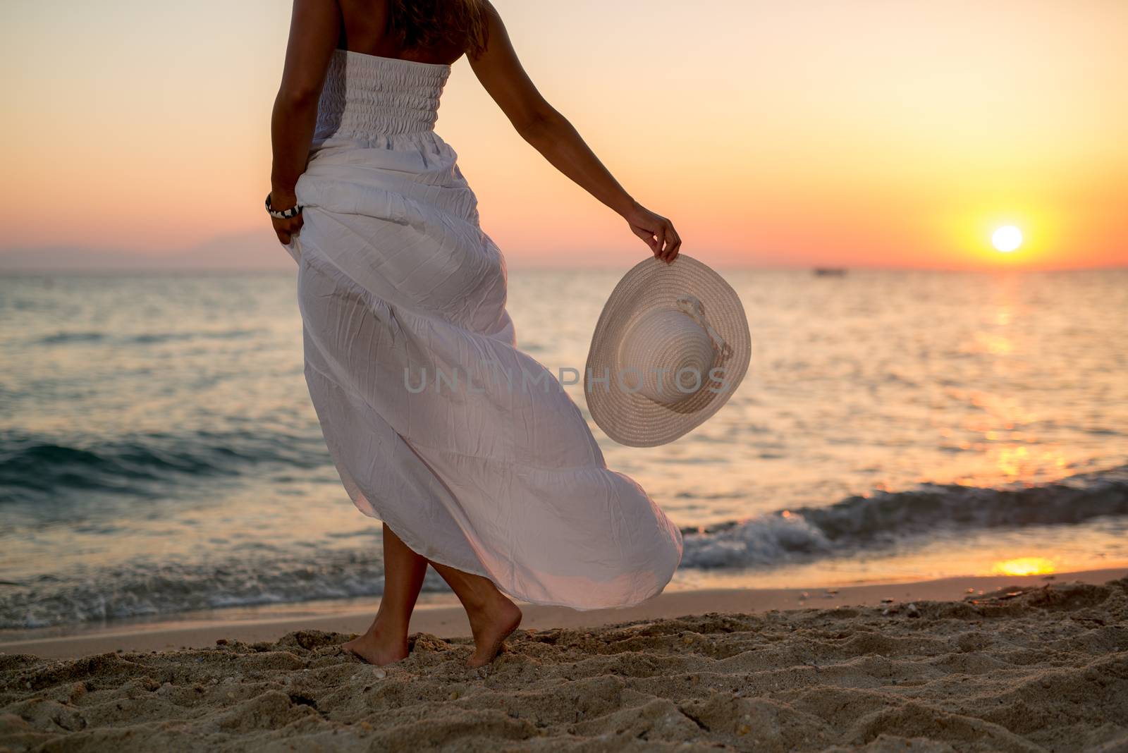 Woman in white dress enjoying sunset on the beach and holding white hat. Rear view.