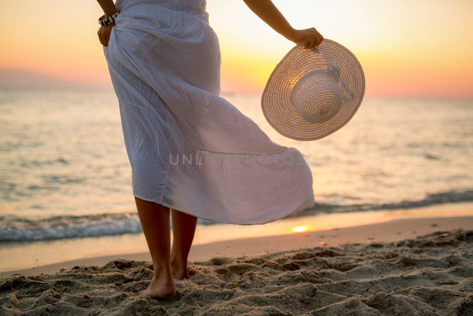 Woman in white dress enjoying sunset on the beach and holding white hat. Rear view.