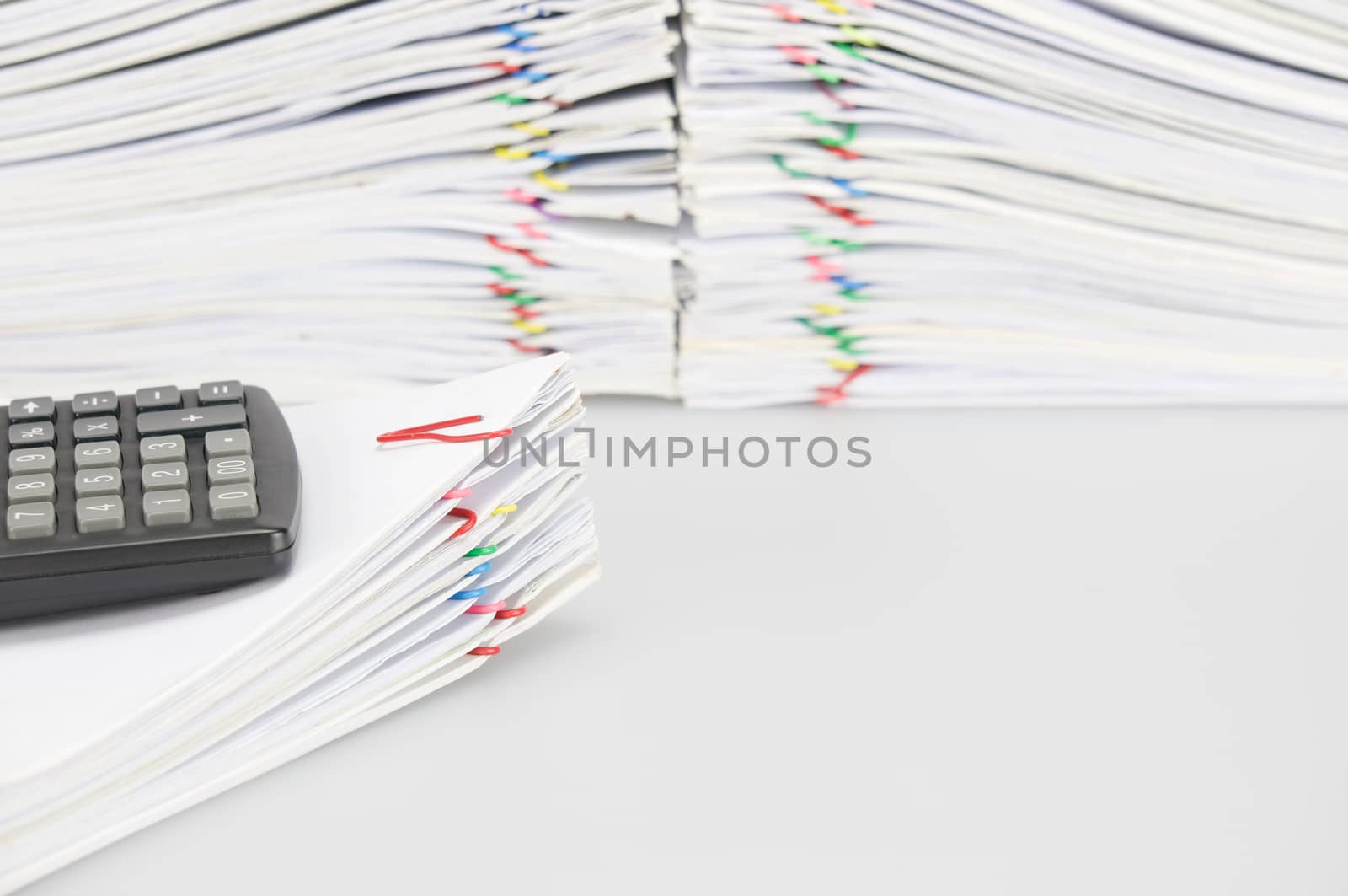 Black calculator place on pile of document have blur overload of paperwork with colorful paperclip on white background.