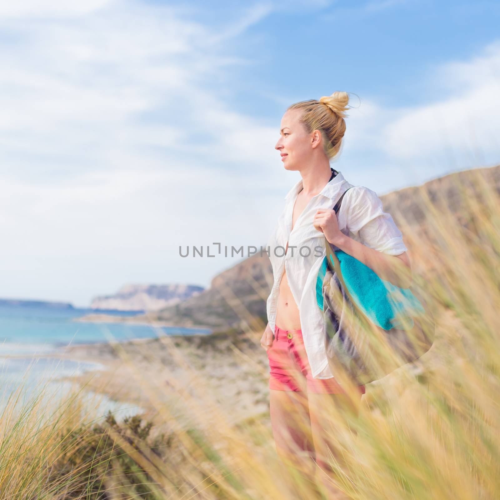 Relaxed woman on vacations in white loose shirt carrying beach bag and towel, enjoying beautiful coastline view of white sandy lagoon at Balos beach, Greece.