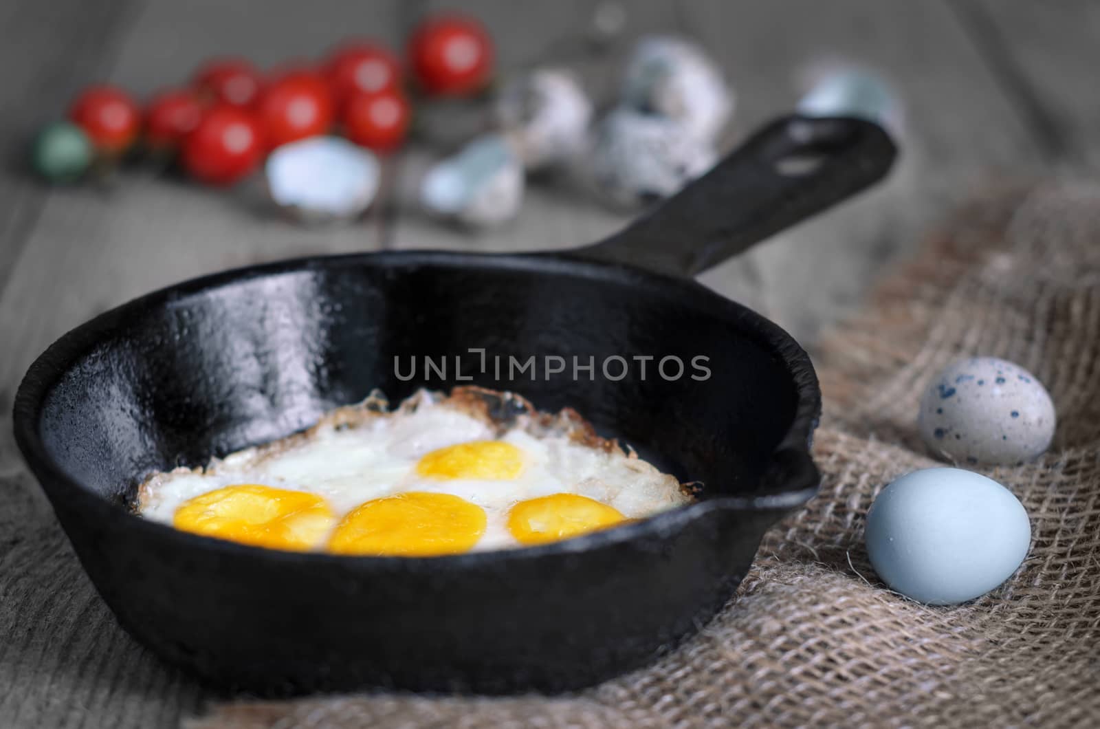 Fried quail eggs on a cast iron skillet, the old wooden background. Small tomatoes and whole eggs. The rustic style.