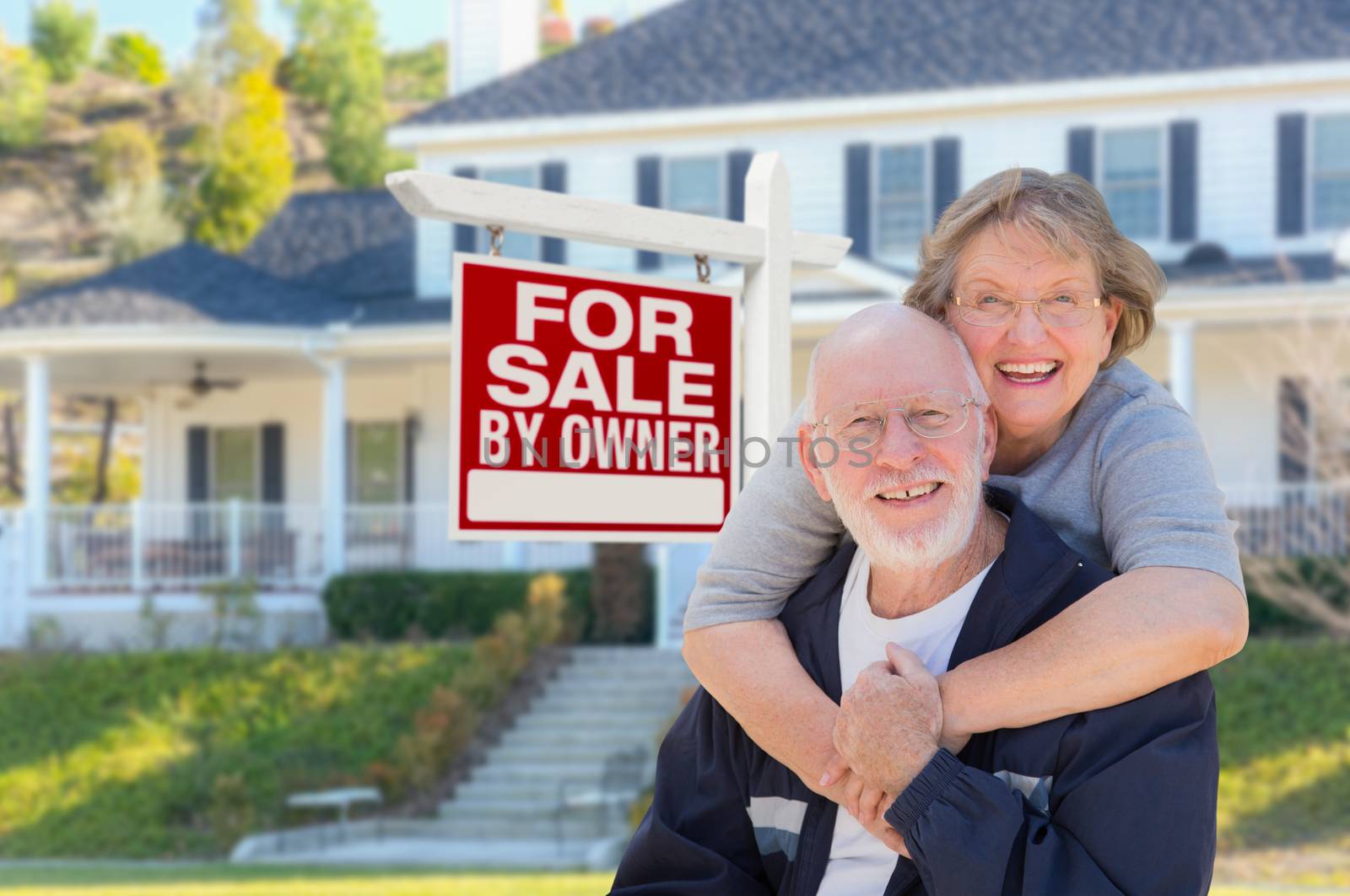 Senior Adult Couple in Front of Home For Sale Real Estate Sign and Beautiful House.
