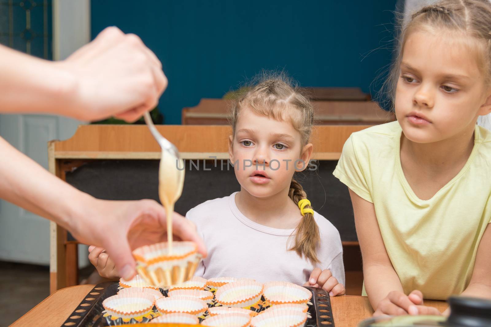Two girls with a surprised look like mom pours batter into molds for cupcakes