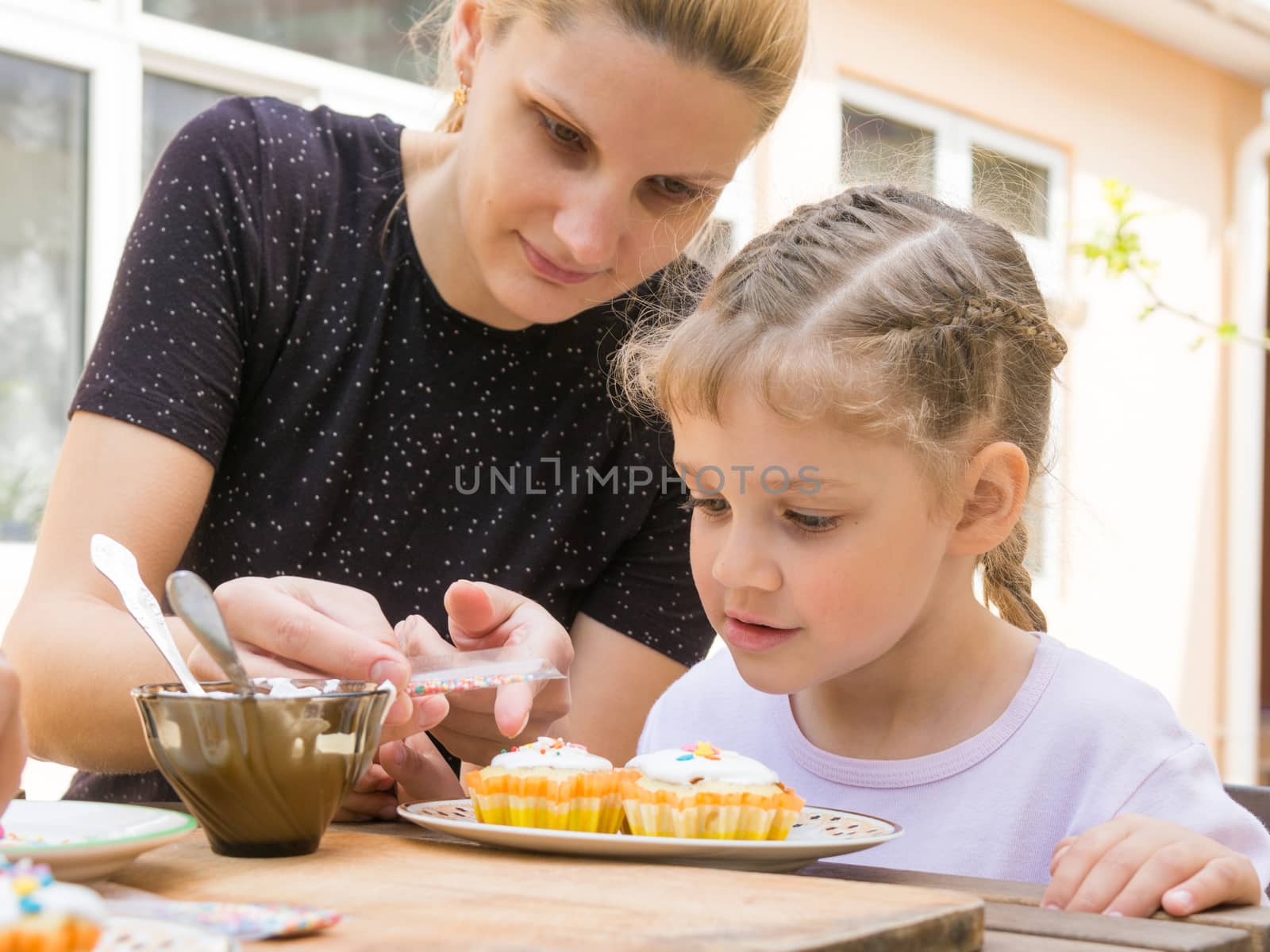 Mother pours a bag of confectionery posypku on Easter cupcake, daughter happily looking
