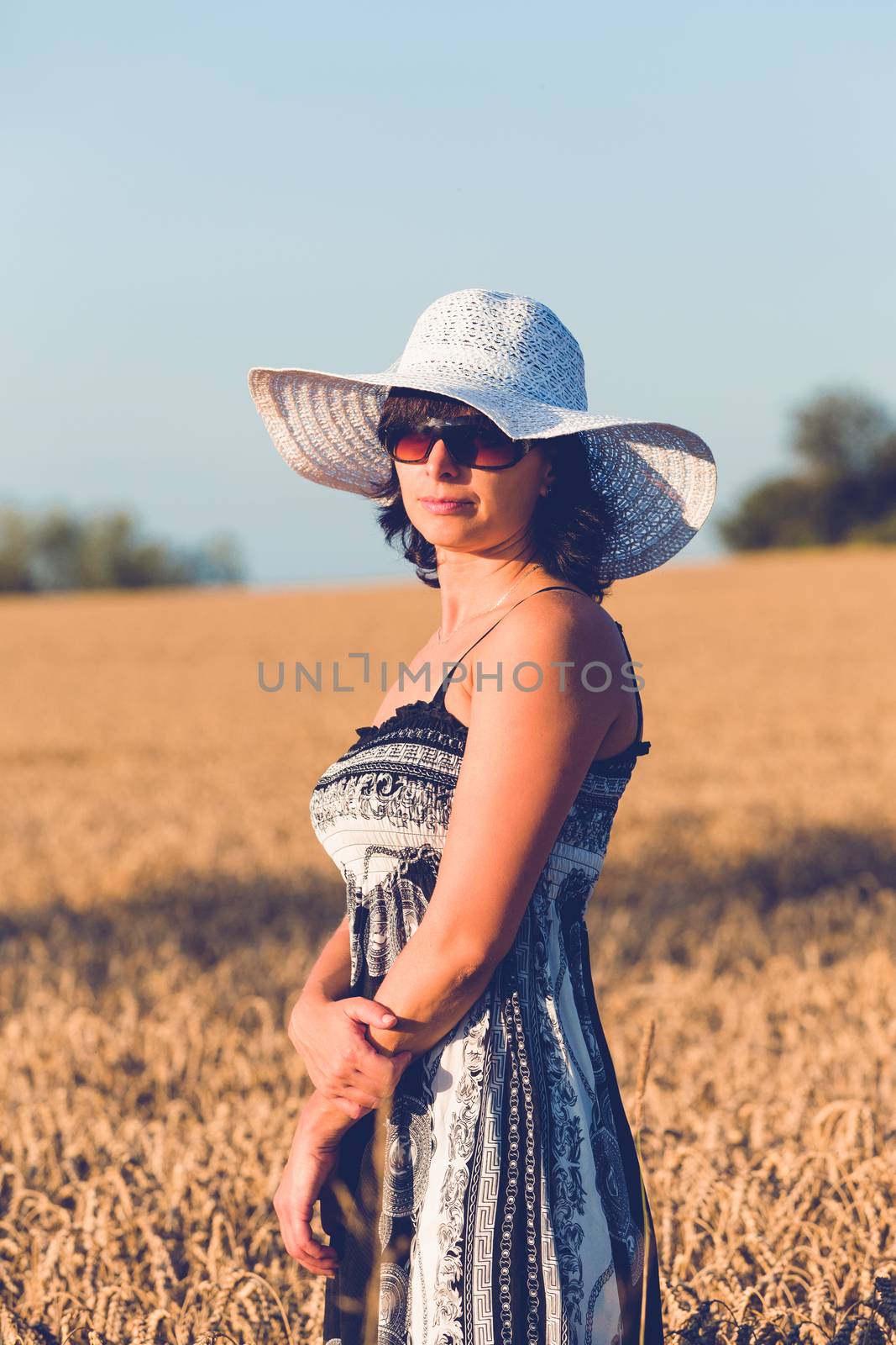 Middle aged beauty woman in a summer dress with summer hat relaxing in countryside, summer concept with evening sun