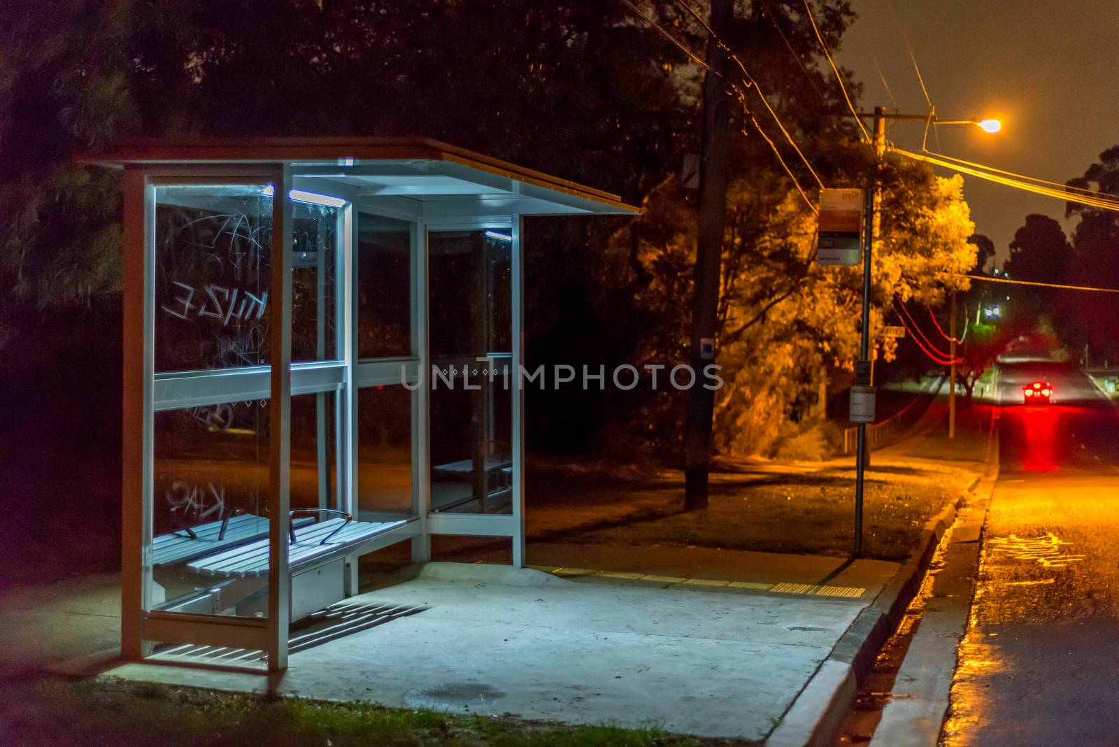 Empty bus shelter at night.  A car is dissappearing down the road.  graffiti is scratched into the glass.