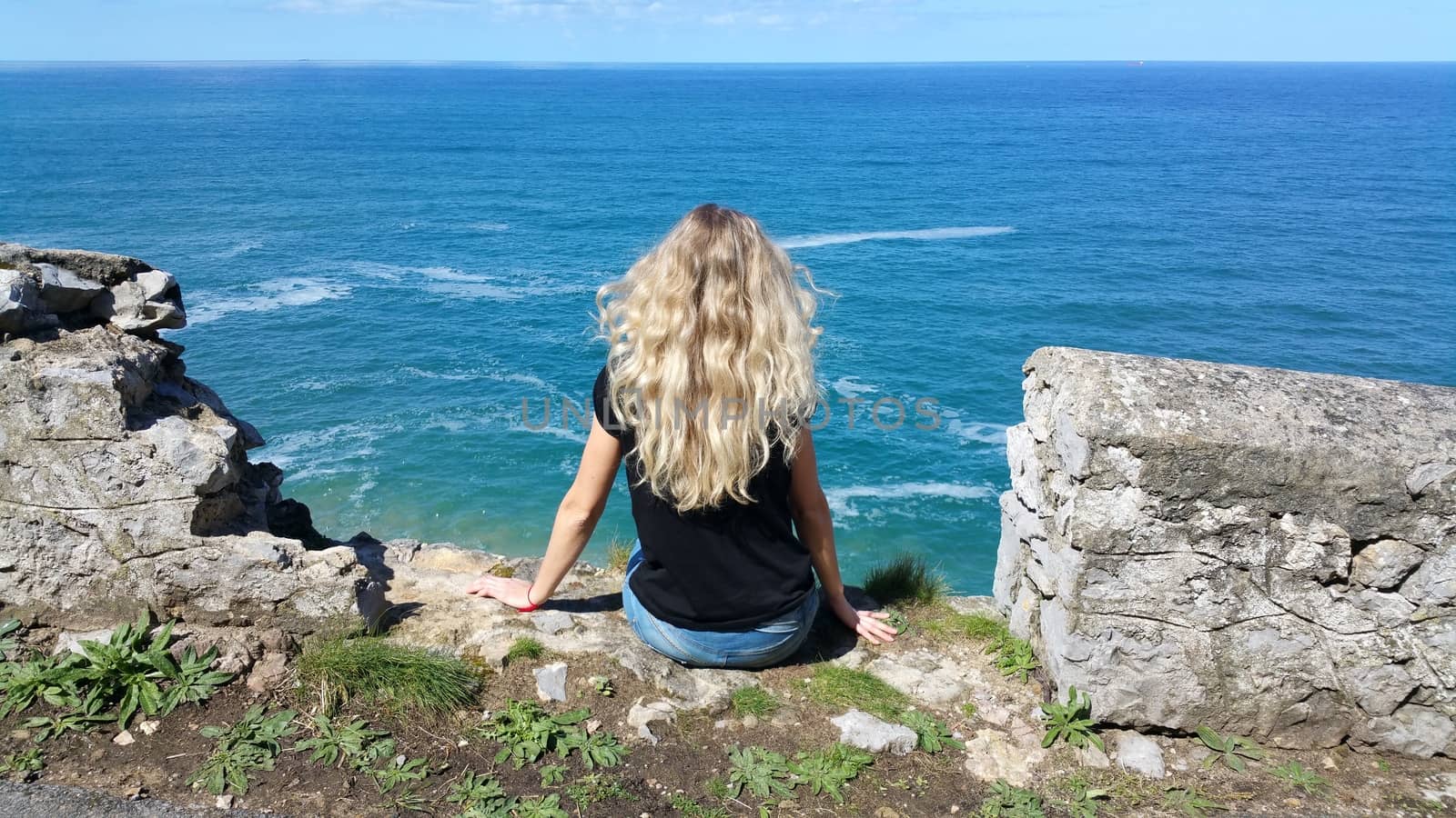 blonde girl sitting back on the rock, looking on the water