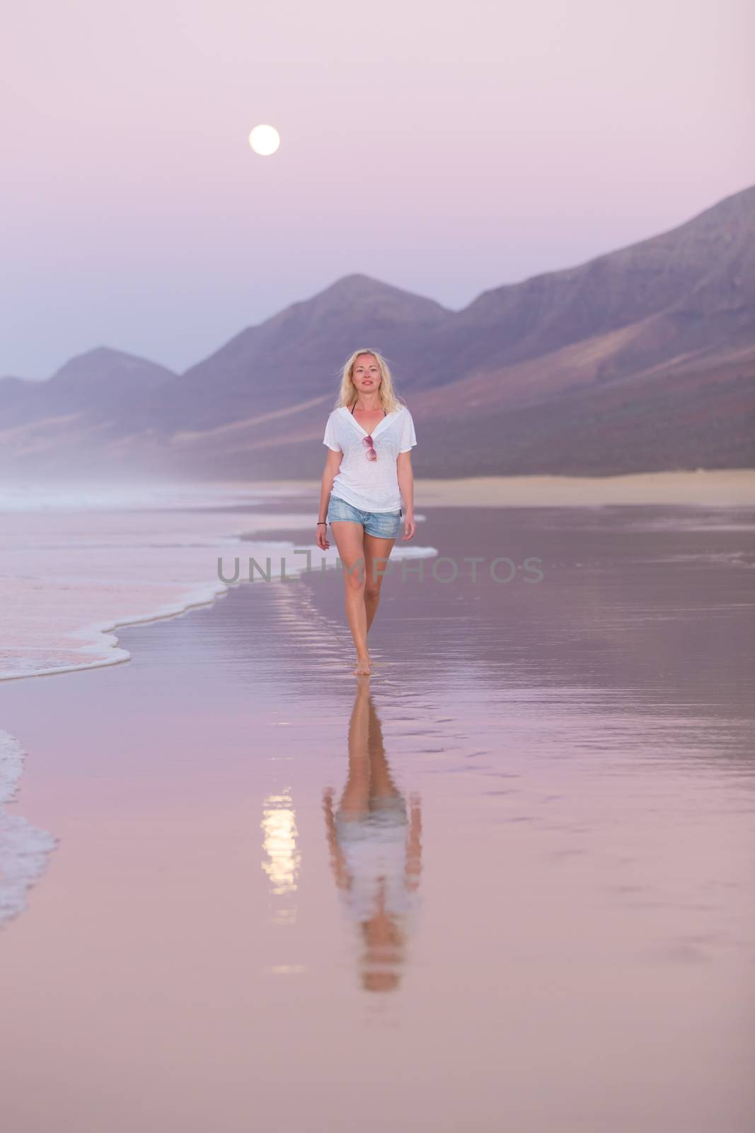 Woman walking on sandy beach in dusk leaving footprints in the sand. Beach, travel, concept. Copy space. Vertical composition.