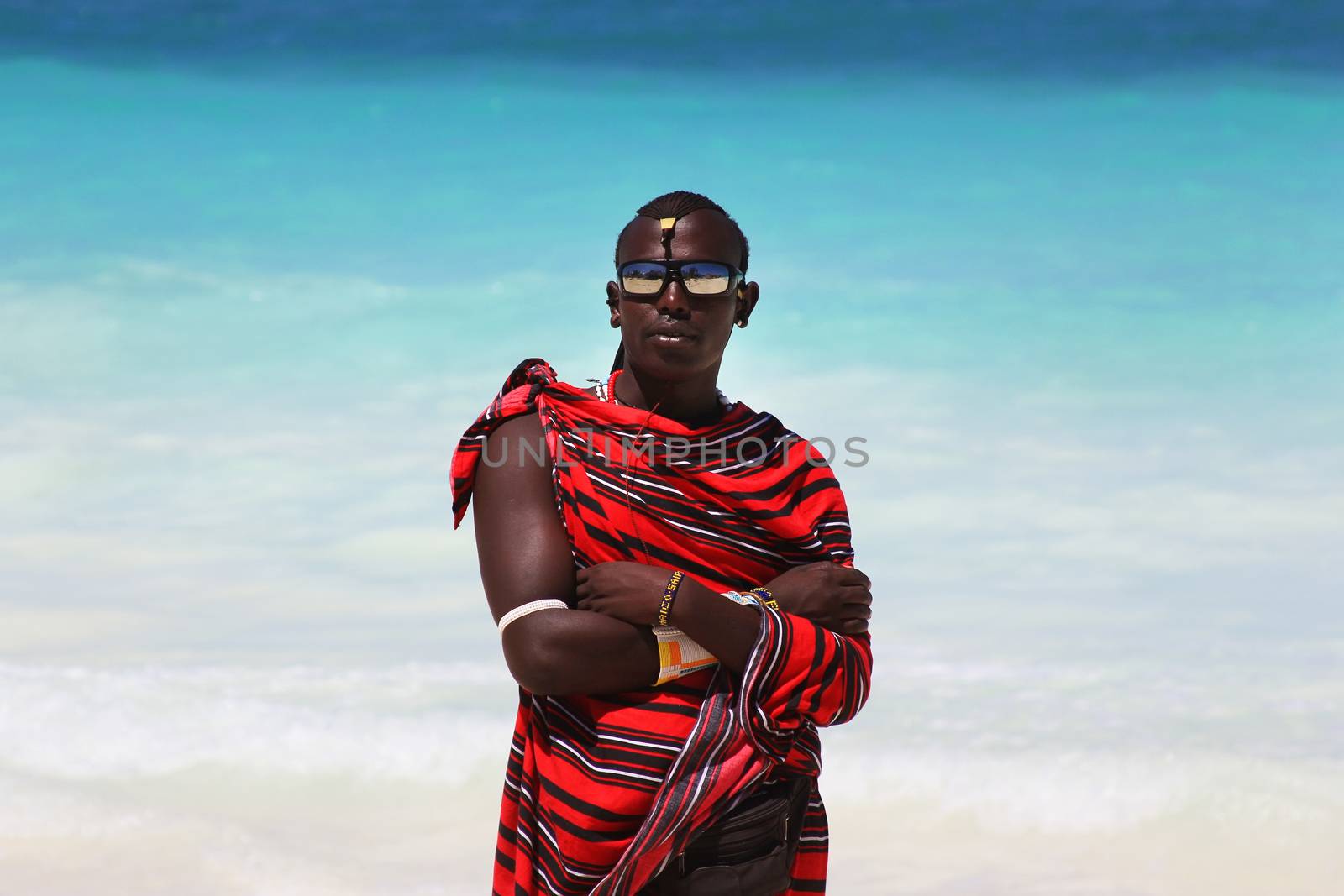 Zanzibar, Tanzania - January 5, 2016: Young massai man with colorful traditional clothes, headdress and self made shoes walking through the water on the Nungwi beach.