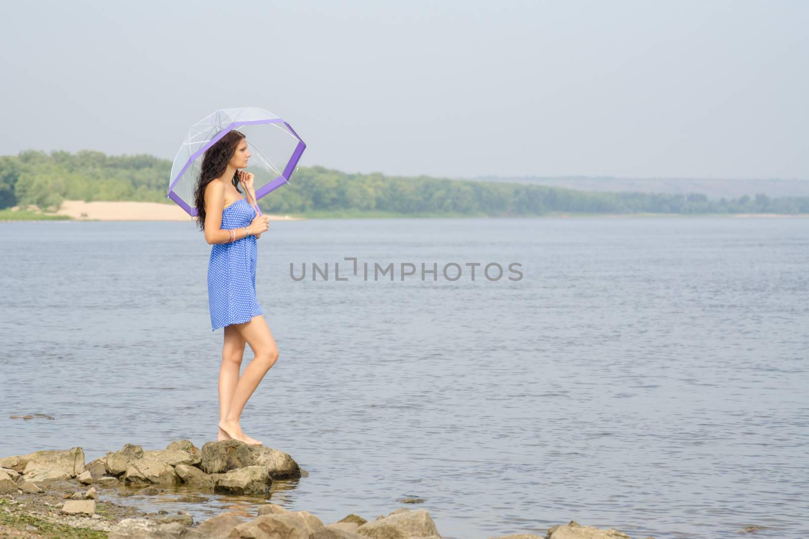 Lonely sad young girl with an umbrella stands on the bank of the river and looks into the distance