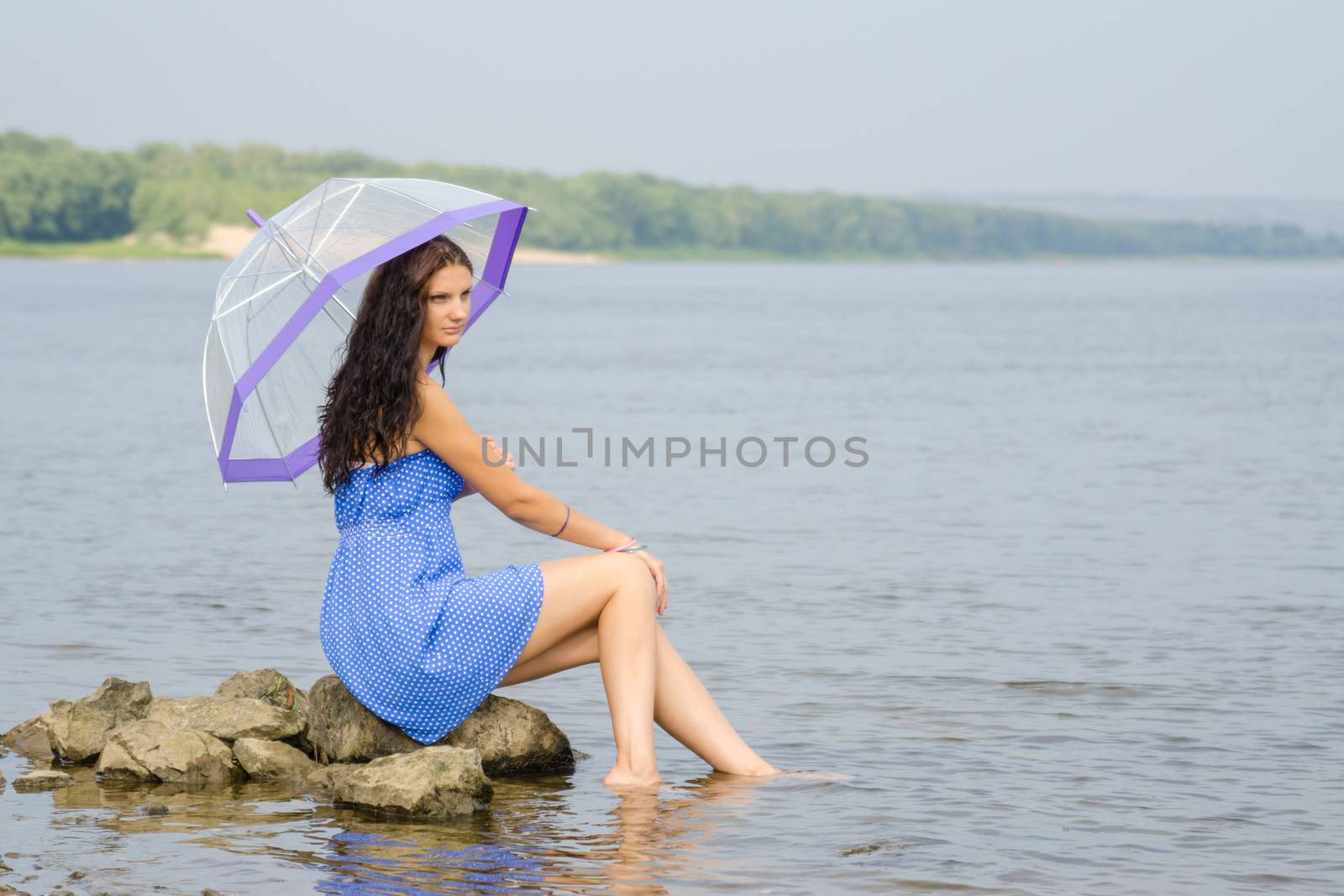 Lonely sad young woman with an umbrella sits on a rock by the river by Madhourse