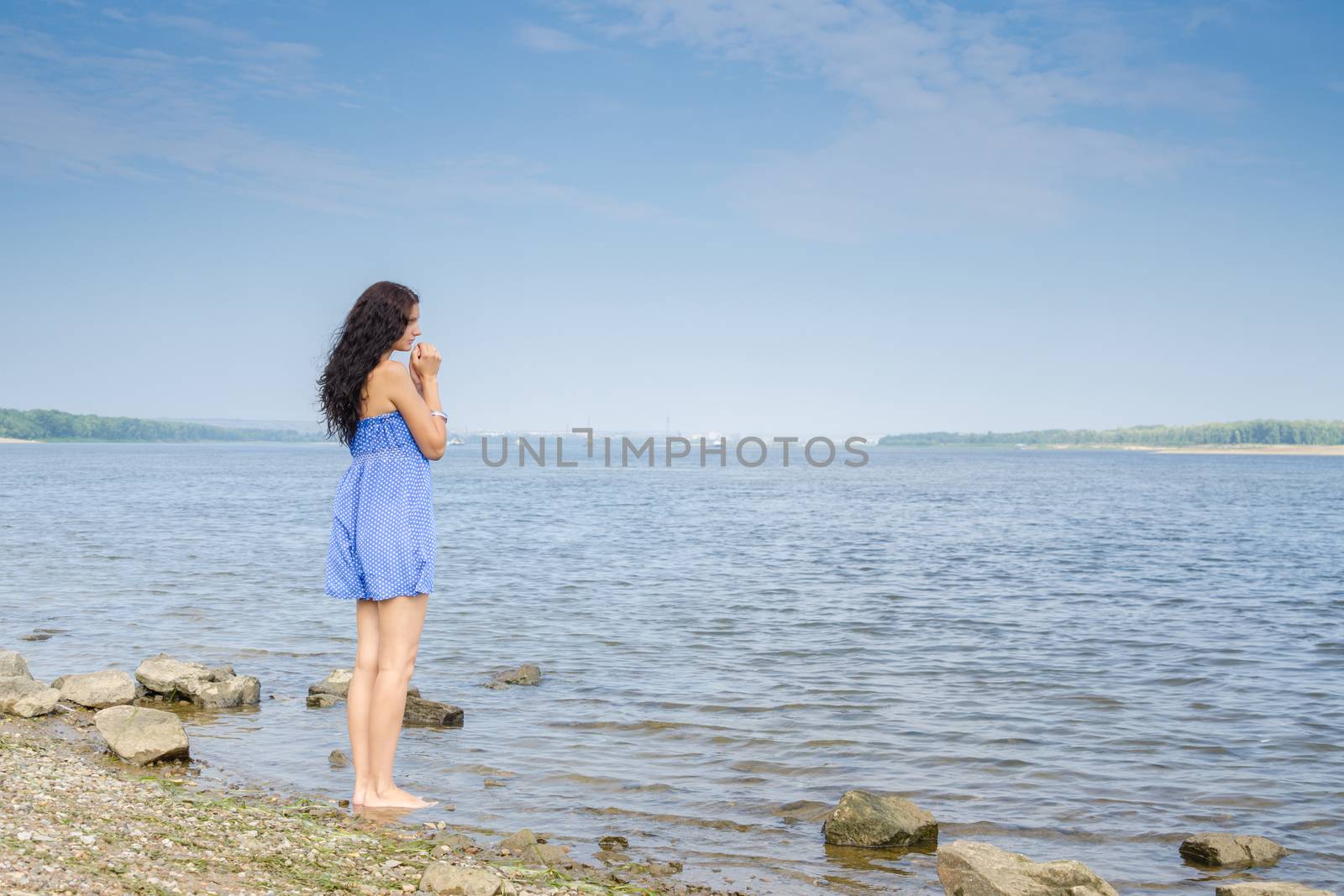 Sad young brunette woman in a blue summer dress standing on the river bank