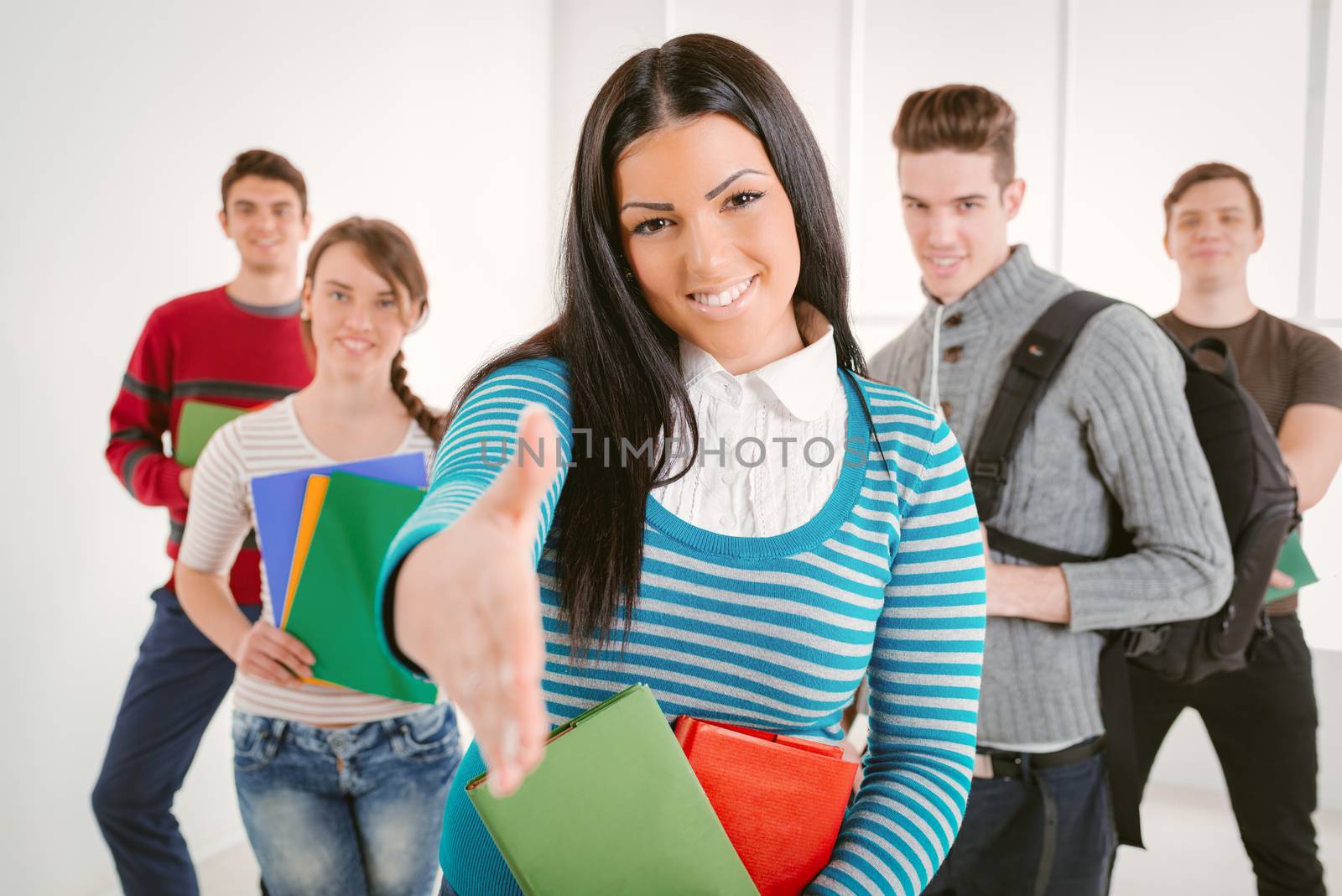 Beautiful smiling girl with books standing in school hall and offering her hand as if to shake hands. Happy group of her friends standing is behind her and looking at camera. Selective focus.