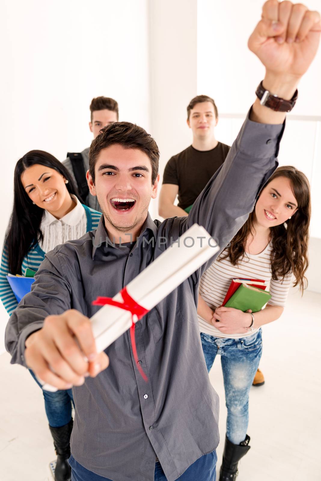 Beautiful successful smiling student with diploma standing in school hall with arms raised in a fist. Happy group of his friends standing is behind him and looking at camera. Selective focus. 